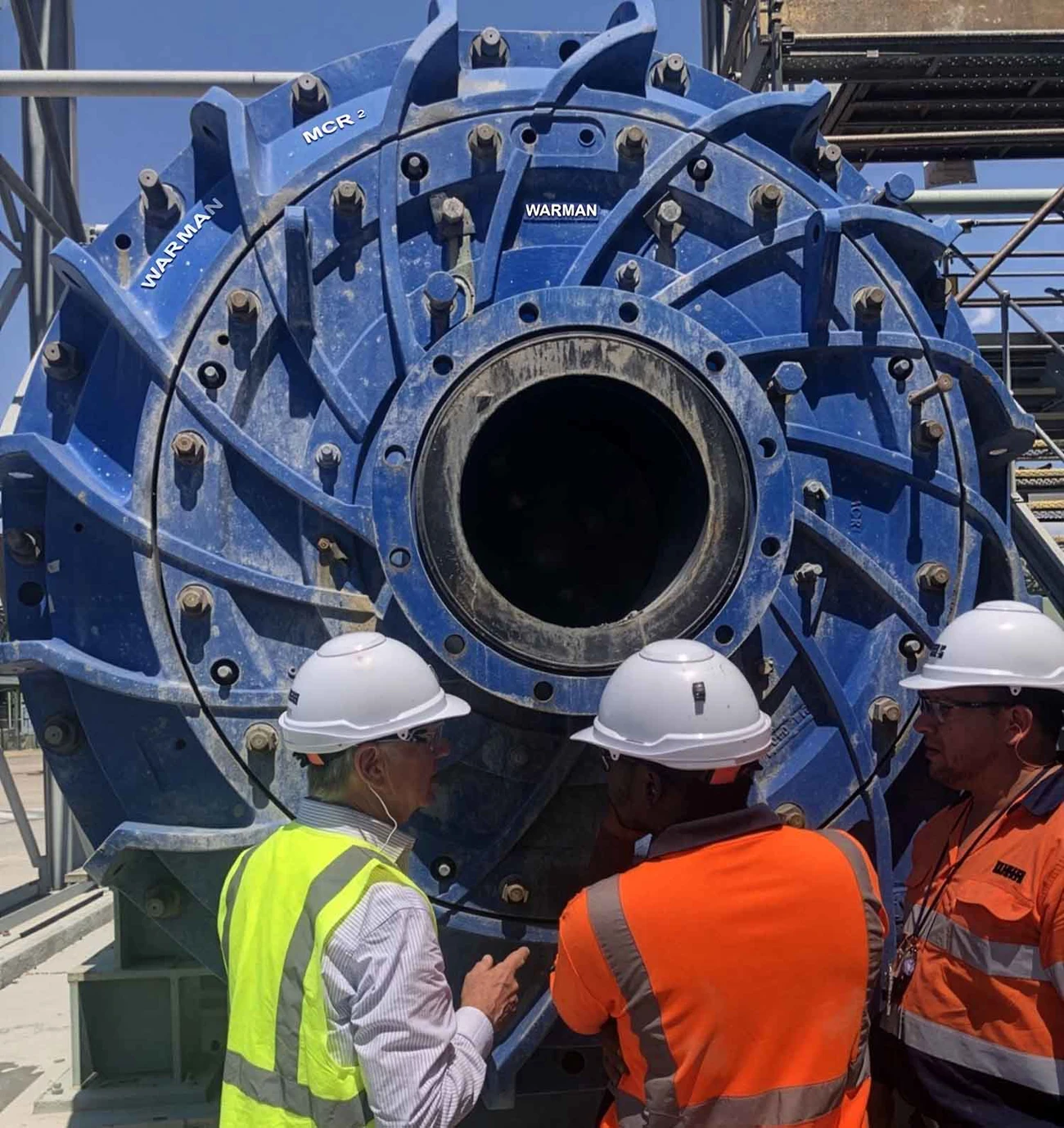 Large blue WARMAN® MCR²® slurry pump installed at an industrial site, with three workers wearing high-visibility safety clothing and white hard hats standing in front of the pump for scale.